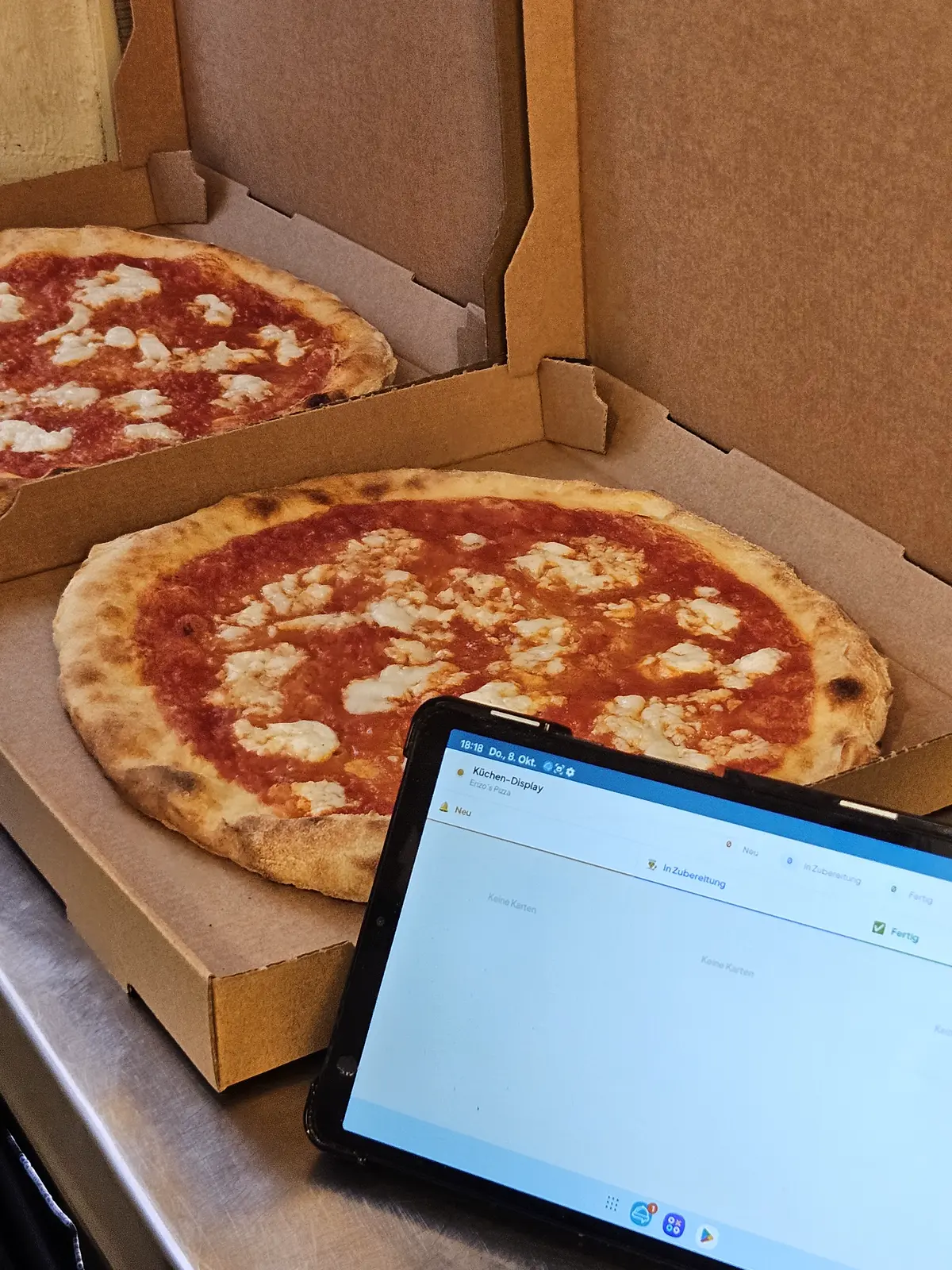 Zwei Pizzen im Karton, davor das Tablet mit der Küchenansicht